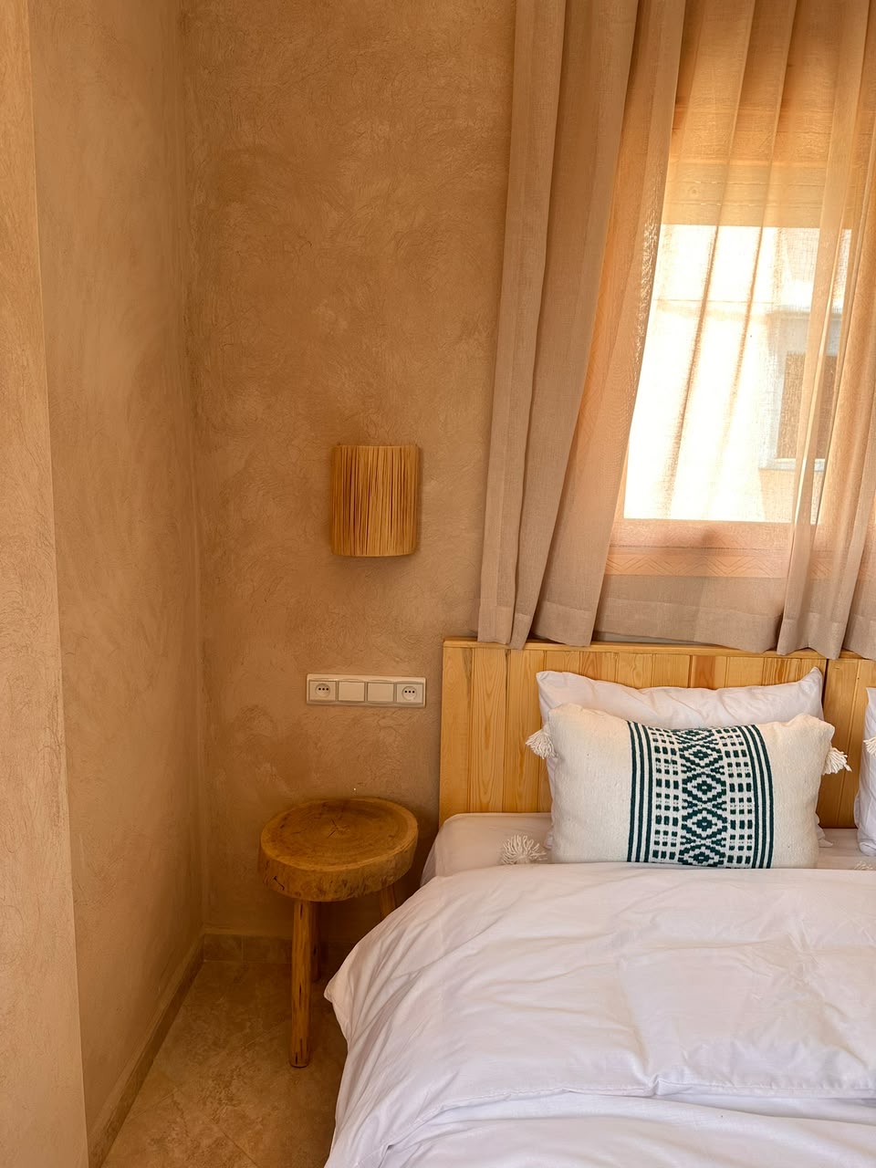Bedside corner with linen curtains, wall lamp and a wooden stool